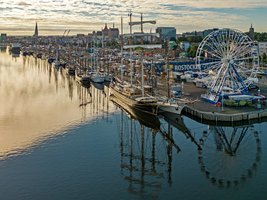 Viele Segelschiffe die an Kaikante liegen, im Hintergrund Jahrmarktbuden und Riesenrad