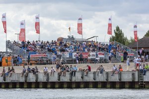 Im Vordergrund Menschen die an der Kai Kante sitzen. Im Hintergrund Tribüne mit vielen Menschen. Auf vielen Flanen steht Wiro.