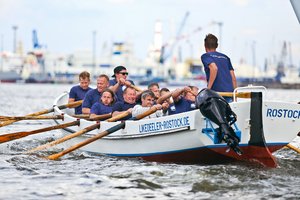 Ein Team von Ruderern im einheitlichen blauen T-Shirts rudert kräftig in einem weiß-blauen Boot mit der Aufschrift "LIKEDEELER-ROSTOCK.DE". Der Steuermann steht hinten im Boot und gibt Anweisungen, während im Hintergrund der Hafen mit Kränen und Schiffen zu sehen ist.
