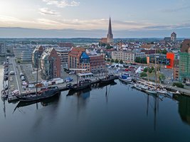 Luftaufnahme von der Sielohalbinsel mit Segelschiffen an Kaikanten und Jahrmarktständen vor den Backsteinsilos. Im Hintergrund die Altstadt von Rostock mit Kirchen  