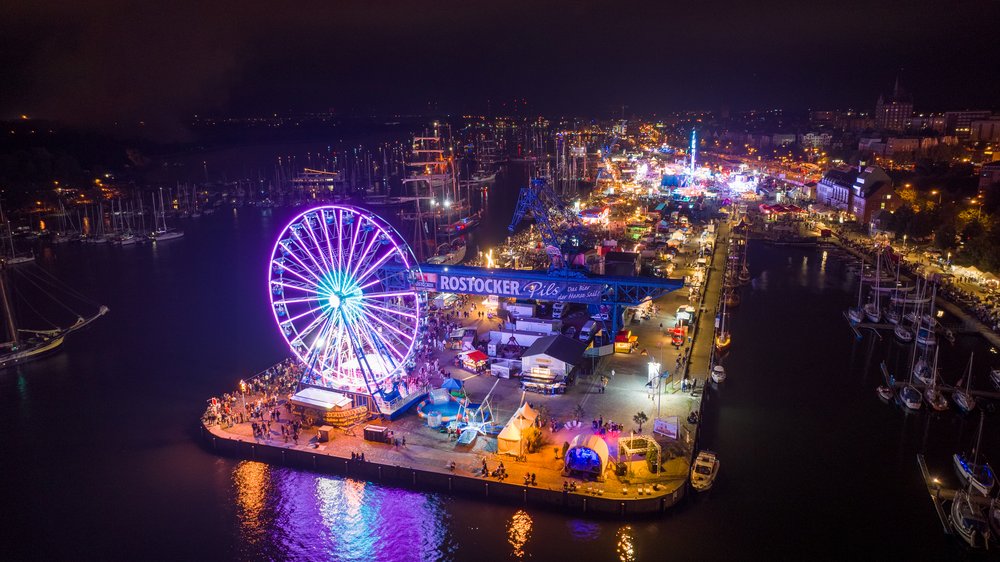 Luftaufnahme der Haedgehalbinsel bei Nacht. Mit Riesenrad, Festzelten und Segelschiffen auf der Warnow