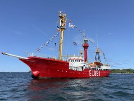 Schiff mit roten Rumpf, weißen Aufbauten und gelben Masten mit bunten Flaggen. In der Mitte hat das Schiff einen Leuchtturm  